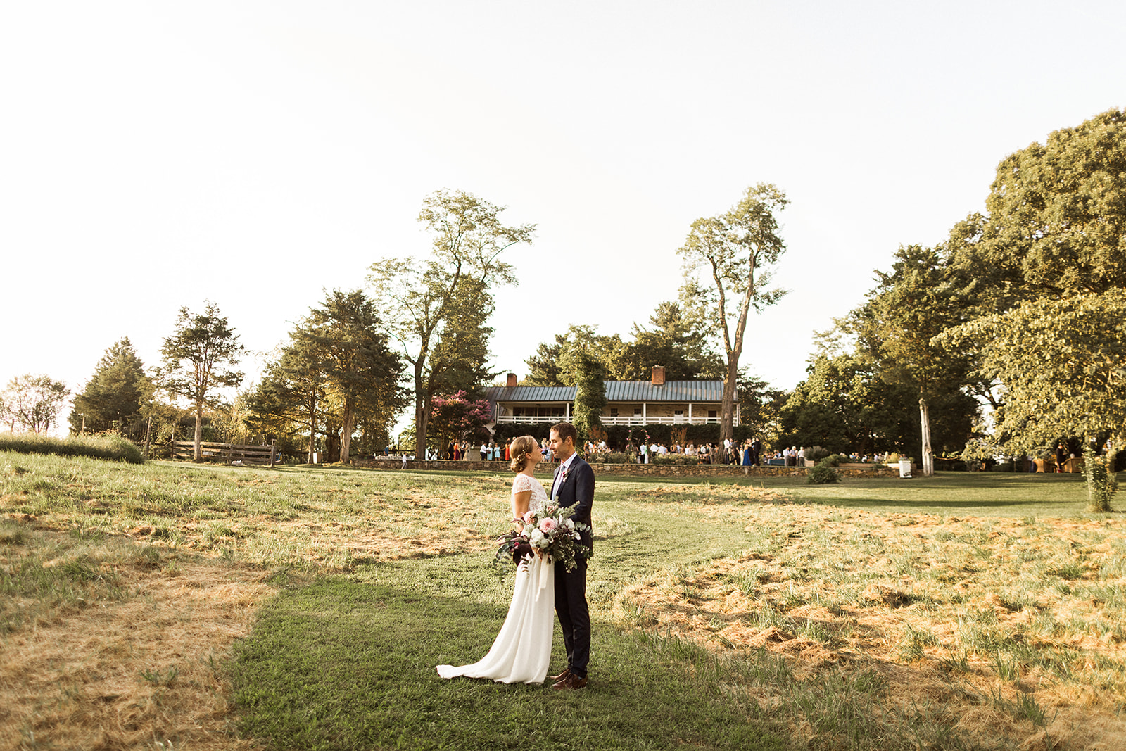 Bride and groom standing in field with guests and farmhouse behind them. 