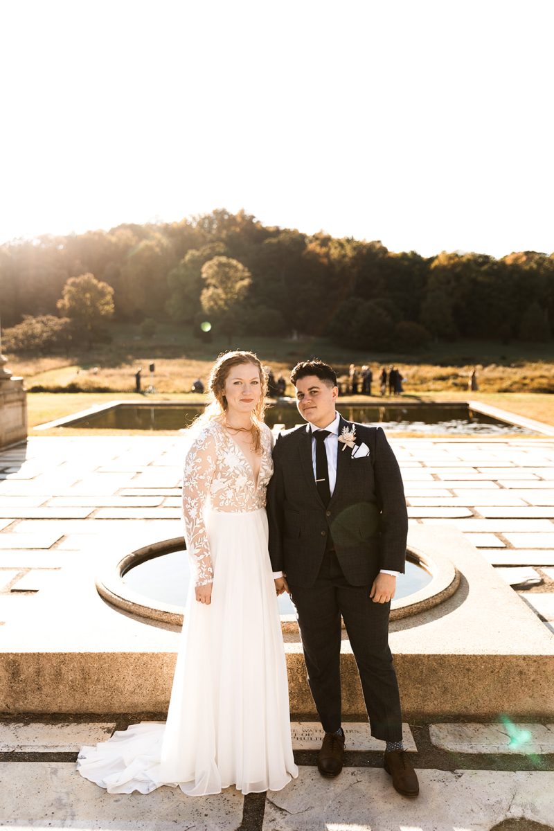 LGBTQ+ couple looking at camera, standing in front of water feature at National Arboretum. 
