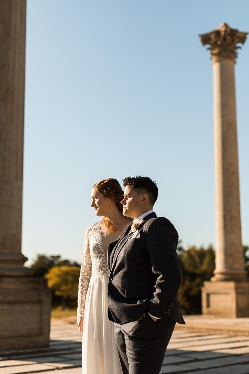 LGBTQ+ couple in wedding attire looking to left of frame in front of columns at National Arboretum. 