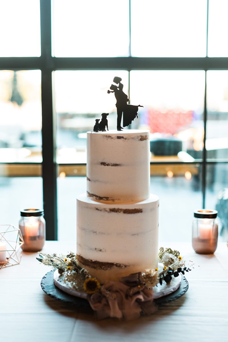 Two tier wedding cake with dried flowers around base and silhouette cake topper featuring two brides and two dogs. 
