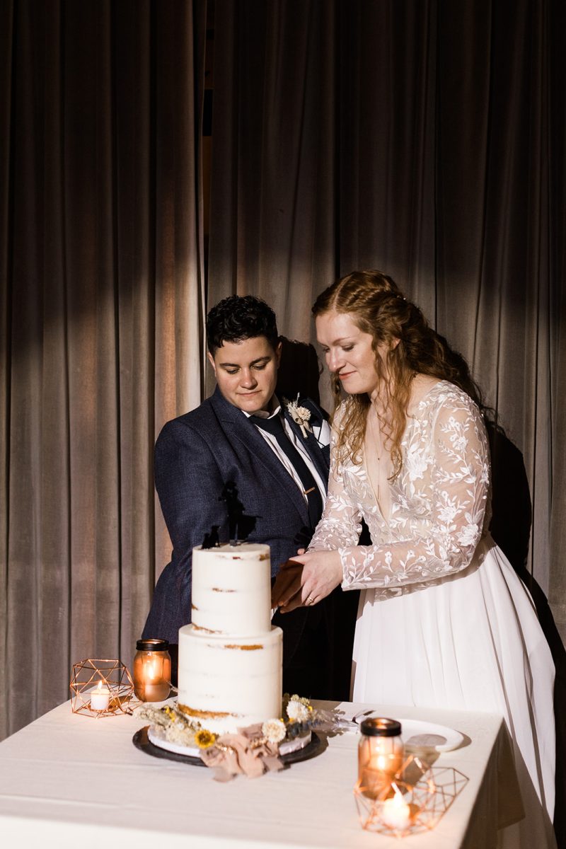 LGBTQ+ couple wearing wedding attire cutting cake. 