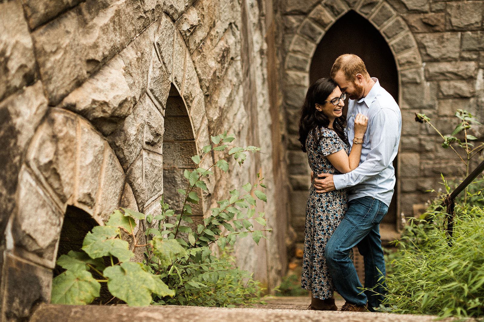 couple embracing standing against stone walls with arched windows and greenery