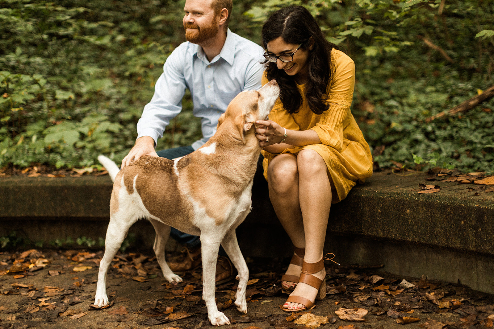 man and woman petting tan and white dog while sitting on curb in forest