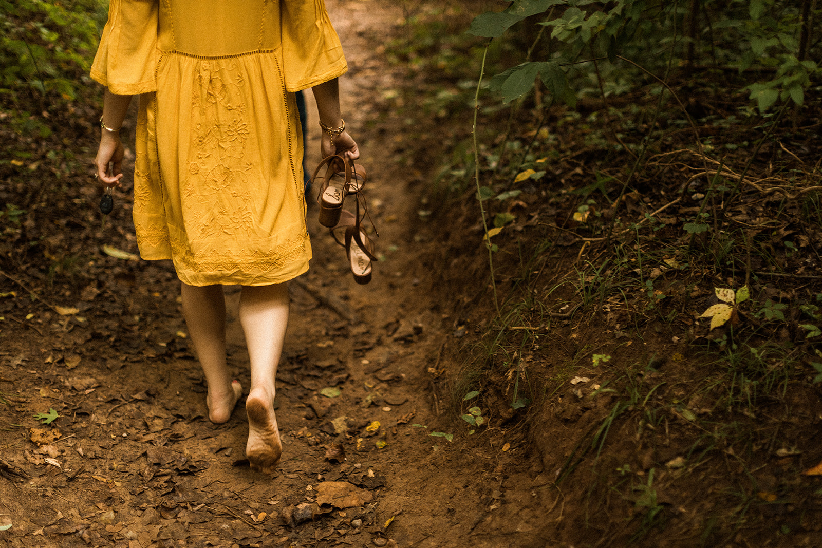 woman in yellow dress walking through forest barefoot while holding high heel shoes near james river