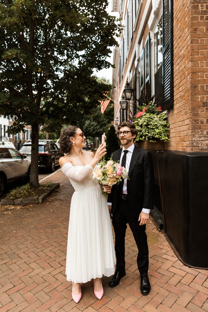 bride holding up small electric fan for groom as they walk along the sidewalk in alexandria virginia