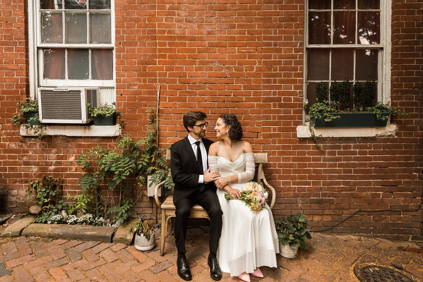 bride and grom sitting on small bench in front of brick wall in alexandria, virginia