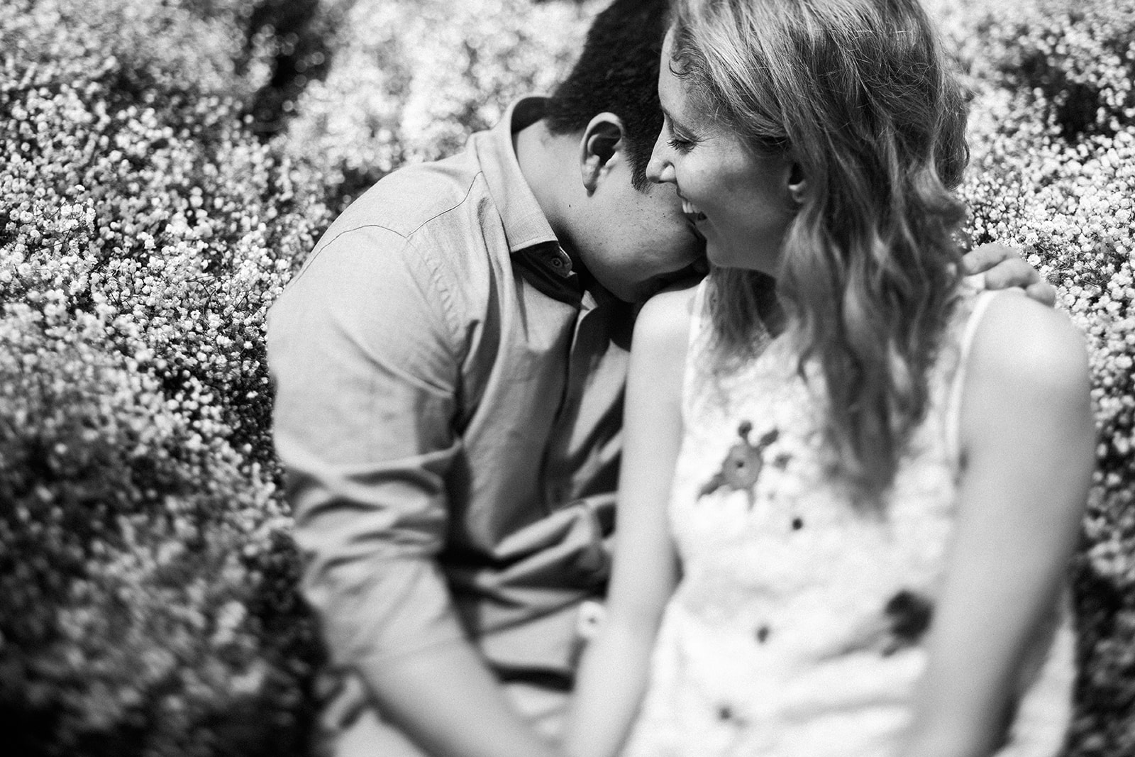 man kissing woman's shoulder in front of flowering bush