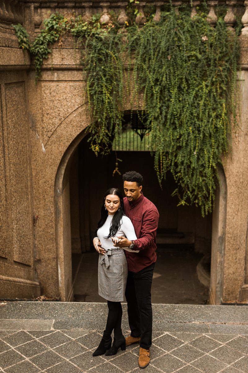 man and woman standing in front of arch in meridian hill park looking at engagement ring
